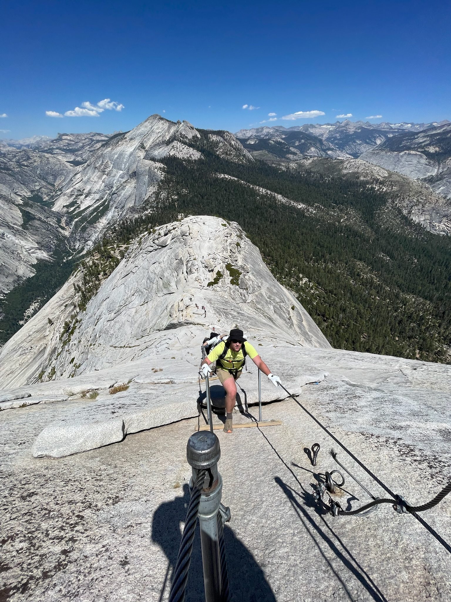 A hiker in a yellow shirt pulling up the steel cables on Half Dome's granite, with forested ridges and the High Sierra behind.