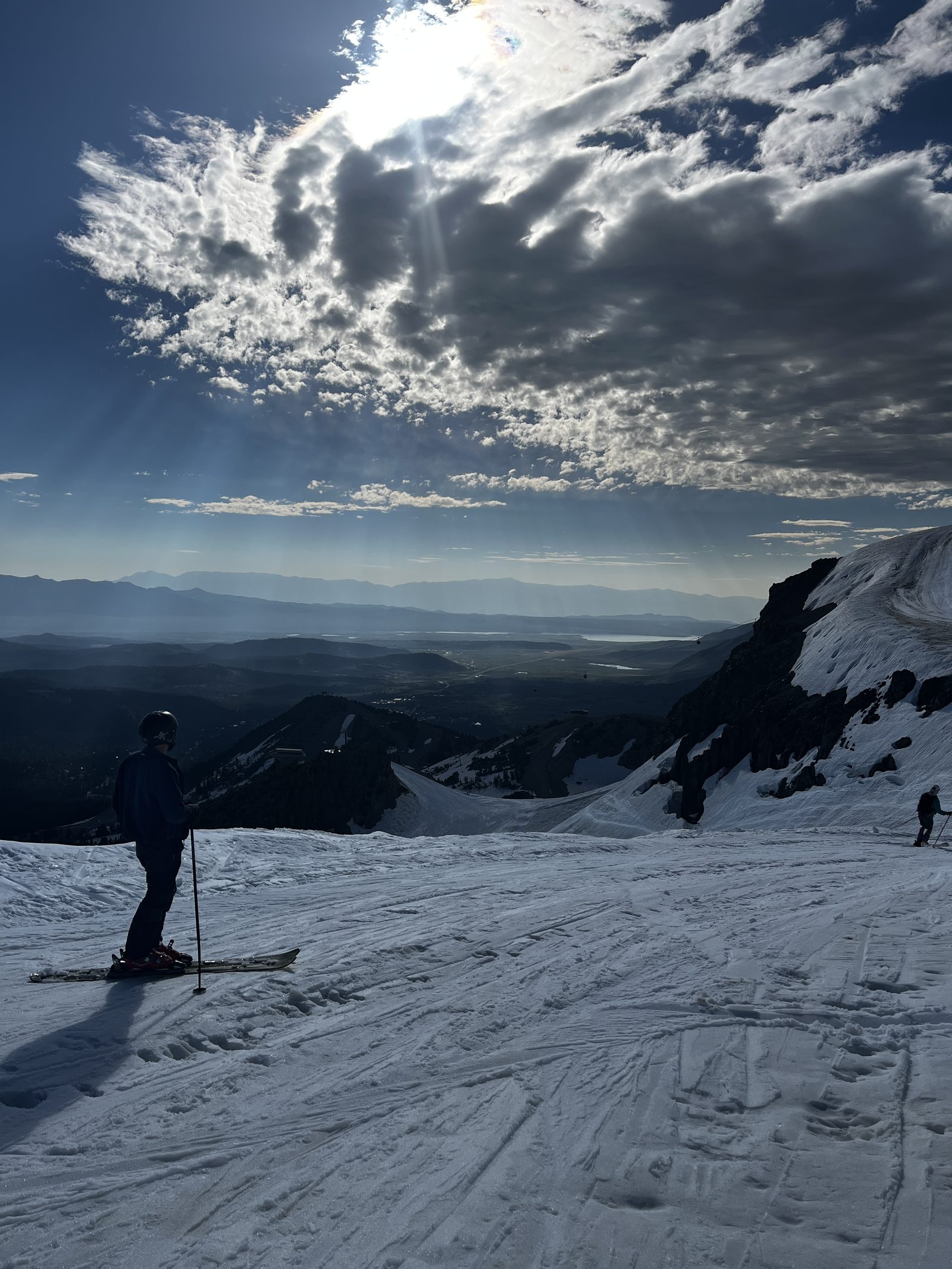 A skier silhouetted on a snowy summit slope, looking out over the Owens Valley under sunlit clouds.